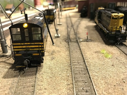 A model train engine on a track with another train in the background, set in a detailed miniature landscape.