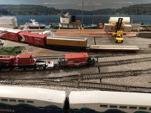 A model train setup featuring freight cars, a tugboat, and a scenic waterfront background with trees and a dock.