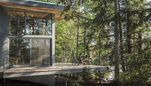 A modern house with large windows, surrounded by trees, featuring a deck where a person relaxes in a chair.