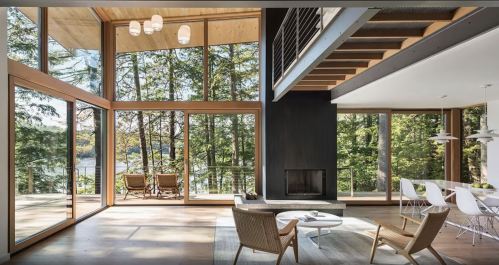 Modern interior with large windows, wooden beams, and a view of trees and water. Cozy seating area and dining space.
