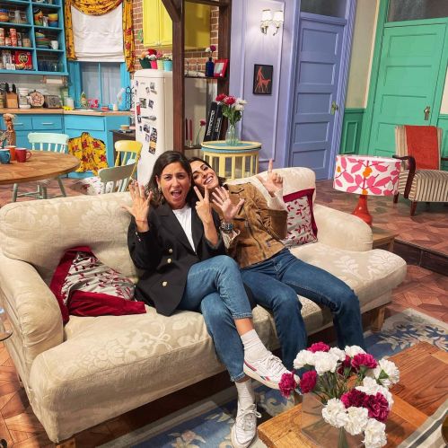 Two people sitting on a couch in a colorful living room, making playful hand gestures and smiling at the camera.