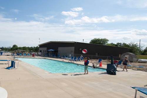 A child throws a beach ball near a swimming pool with lounge chairs and a building in the background on a sunny day.