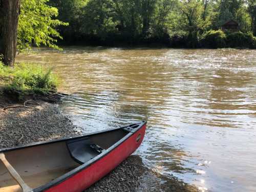 A red canoe rests on a rocky shore beside a calm, muddy river surrounded by lush greenery.