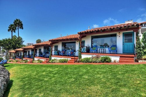 Colorful beach cottages with blue doors and chairs, surrounded by green grass and palm trees under a clear blue sky.