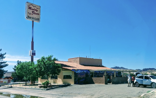 A rustic bar and grill building with a sign, surrounded by trees and a clear blue sky. A car and people are nearby.