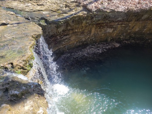 A small waterfall cascades into a deep, clear pool surrounded by rocky terrain and dry leaves.