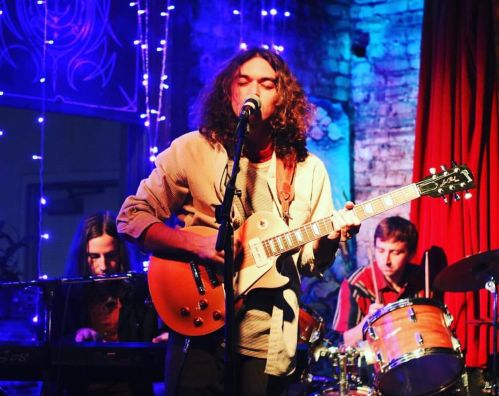 A musician performs with a guitar on stage, accompanied by a pianist and drummer, with colorful lights in the background.
