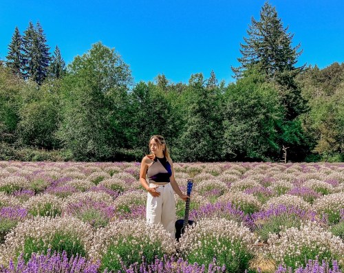 A woman stands in a lavender field, surrounded by blooming flowers and greenery, holding a guitar.