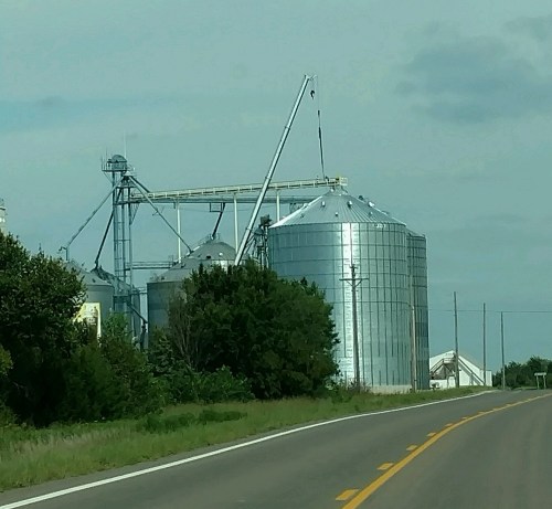 Grain silos and equipment along a rural road, surrounded by greenery under a cloudy sky.
