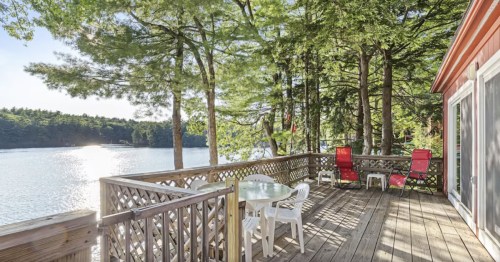 A serene lakeside deck with a table and chairs, surrounded by trees and reflecting water in the background.