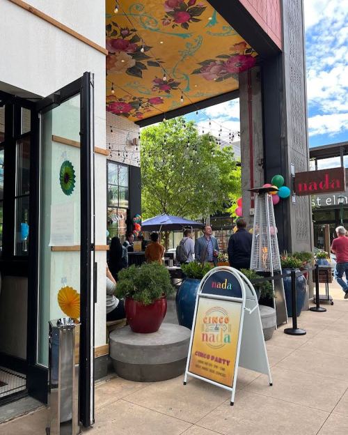 Exterior view of a restaurant with colorful decor, potted plants, and a sign for a Cinco de Mayo party.
