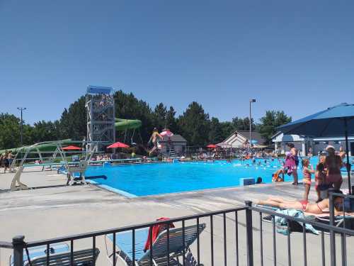 A busy outdoor pool area with a diving board, water slide, and people swimming and relaxing under umbrellas.