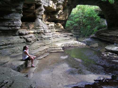 A girl sits on a rock by a serene, wooded creek surrounded by steep, rocky cliffs.