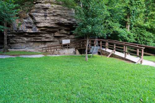 A grassy area with a wooden bridge leading to a rocky cave entrance, surrounded by trees.