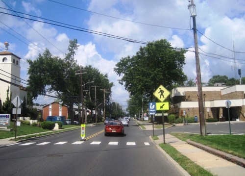 A street view with a red car, pedestrian crossing sign, and trees under a partly cloudy sky.