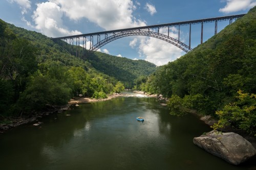 A scenic view of a river surrounded by lush greenery, with a large bridge arching over the landscape under a blue sky.