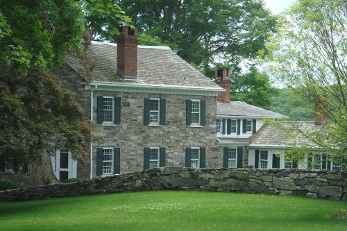 A historic stone house with green shutters, surrounded by lush trees and a stone wall on a sunny day.