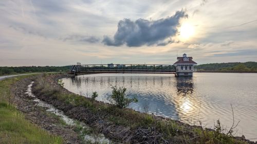 A serene lake scene with a wooden bridge leading to a small building, under a cloudy sky at sunset.
