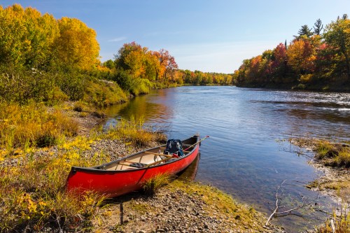 A red canoe rests on a rocky shore beside a calm river, surrounded by vibrant autumn foliage.