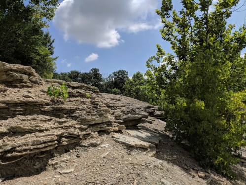 Rocky terrain with layered stone formations, scattered greenery, and a blue sky with fluffy clouds.