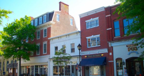 Historic buildings line a sunny street, featuring brick facades and a pub, surrounded by lush green trees.
