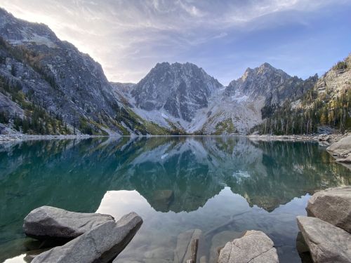 A serene mountain lake reflecting rugged peaks and a clear sky, surrounded by rocky shores and evergreen trees.