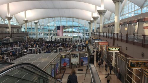 A busy airport terminal with travelers, security checkpoints, and shops under a high, arched ceiling. An American flag is visible.