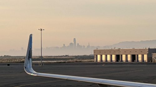 A view of a city skyline shrouded in mist, seen from an airplane wing at an airport.