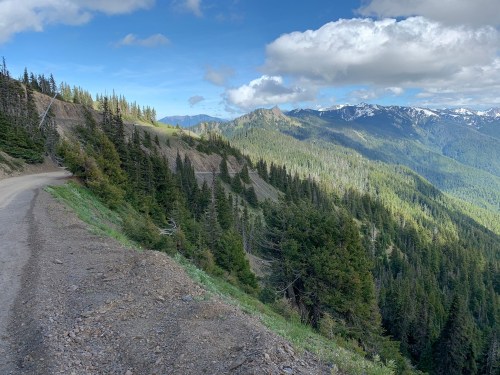 A winding dirt road along a mountain ridge, surrounded by lush green trees and distant snow-capped peaks under a blue sky.