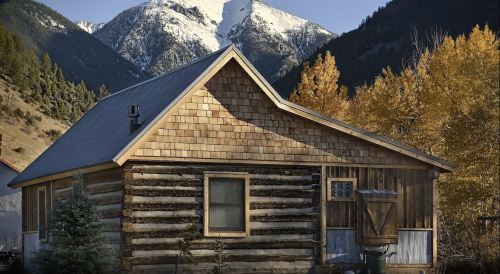 A rustic log cabin surrounded by mountains and autumn trees, with snow-capped peaks in the background.