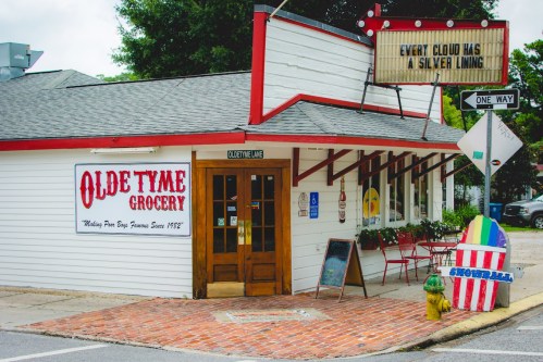 A quaint grocery store with a red and white sign, featuring a vintage design and a colorful surfboard decoration outside.