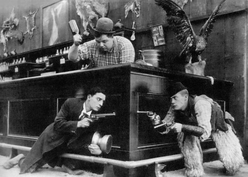 Two men crouch behind a bar, aiming guns, while a third man stands above them, holding a hammer. Vintage black and white scene.