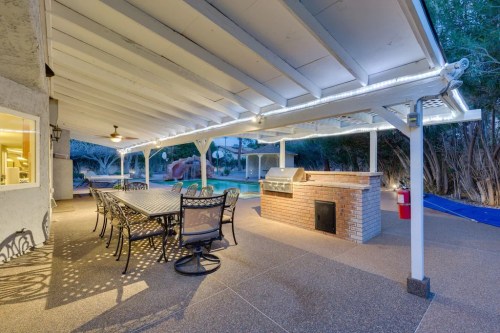 Outdoor patio with a dining table, grill, and ambient lighting, overlooking a pool and surrounded by greenery.