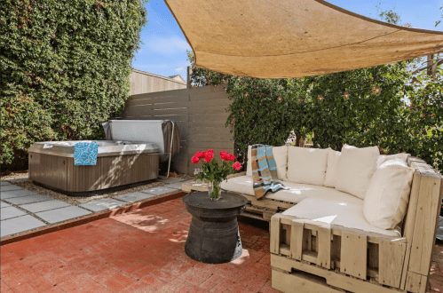 Cozy outdoor space with a hot tub, a pallet sofa, and a table, shaded by a canopy and surrounded by greenery.