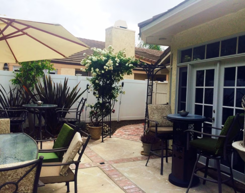 A cozy patio with tables, chairs, and a blooming plant, surrounded by a fence and a cloudy sky.