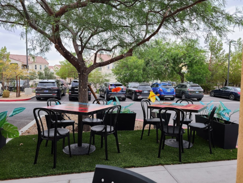 Outdoor seating area with black tables and chairs, surrounded by greenery and parked cars in the background.