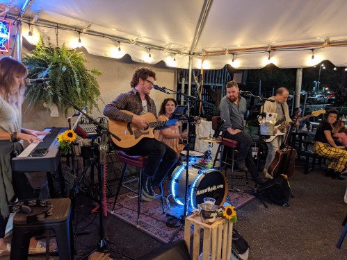 A live band performs in a cozy venue, with musicians playing guitar, piano, and drums, surrounded by plants and decor.