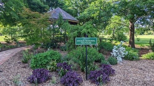 A sign for the Kroze Memorial Tea House surrounded by lush greenery and colorful flowers in a garden setting.