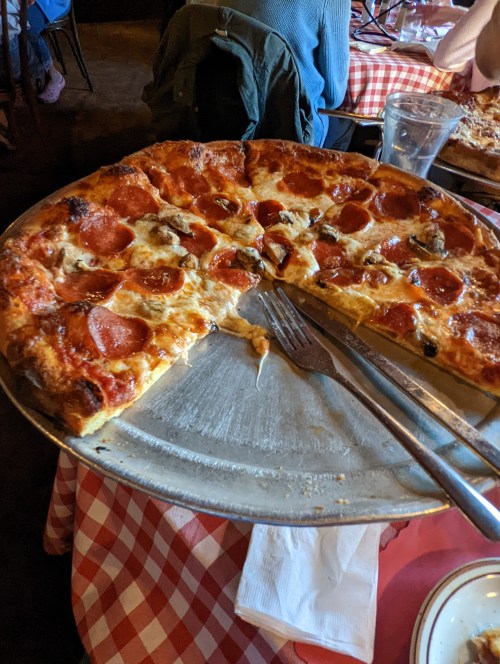 A large pepperoni pizza on a metal tray, with a few slices missing, set on a checkered tablecloth.