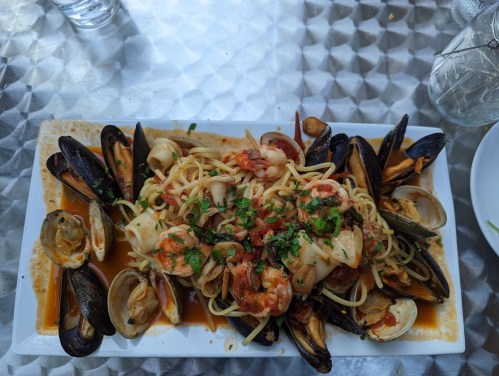 A platter of seafood pasta with shrimp, mussels, and clams, garnished with parsley on a textured silver table.