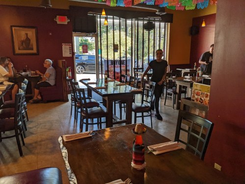 A restaurant interior with tables, chairs, and patrons. A server walks by, and colorful decorations hang from the ceiling.