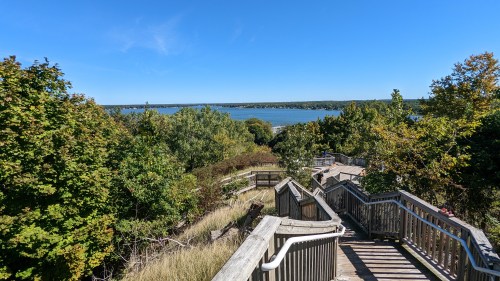 A wooden staircase winds down through lush greenery, leading to a scenic view of a lake under a clear blue sky.