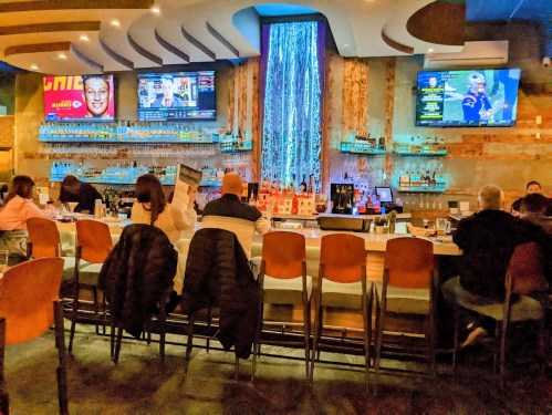 A modern bar with a waterfall feature, illuminated shelves, and patrons seated at the counter watching screens.
