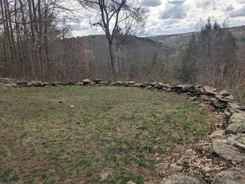 A grassy clearing surrounded by a stone wall, with trees and a distant view of rolling hills under a cloudy sky.