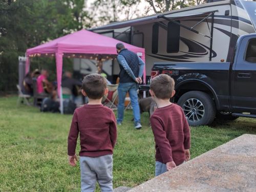 Two young boys in matching outfits stand in a grassy area, watching a gathering near a pink tent and an RV.
