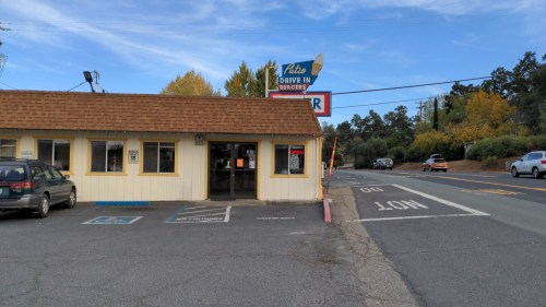 A small drive-in restaurant with a yellow exterior, sign, and parking area, set against a clear blue sky.