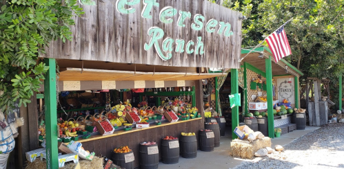 A rustic market stand at Petersen Ranch displays colorful fruits and vegetables, with an American flag nearby.
