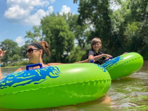 Two people float on green inflatable tubes in a river, smiling and enjoying a sunny day surrounded by trees.