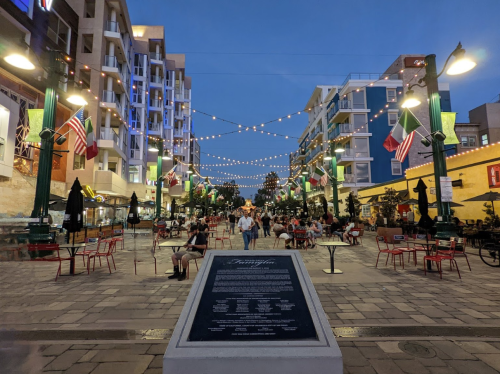 A lively outdoor plaza at dusk, with string lights, flags, and people dining at tables. A historical plaque in the foreground.