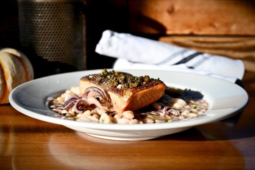 A plated dish featuring salmon with a herb crust, served on a bed of beans and octopus, with a rustic background.